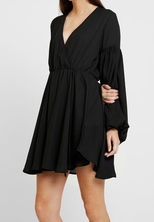 Kobieta ubrana w czarną mini sukienkę z długim rękawem, dekoltem w serek i marszczoną talią, stojąca z jedną zgiętą ręką, której dłoń spoczywa na przeciwległym ramieniu.