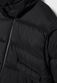 Veste matelassée noire avec coutures diagonales, fermeture éclair à l'avant, poche à rabat et capuche attachée.