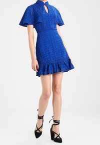 Vestido azul de ojos de perdiz con mangas cortas acampanadas, escote de ojal, cintura entallada y dobladillo con volantes. Combinado con tacones altos negros con tirantes.