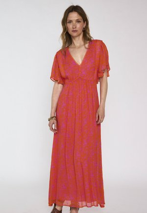 Femme portant une longue robe orange fluide à motif floral rose, col en V, manches à volants, bracelets dorés, debout sur un fond uni.
