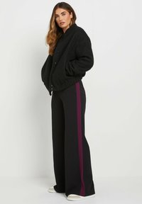Giacca in tessuto nero e soffice con scollatura tonda e bottoni a scatto, abbinata a pantaloni neri a gamba larga con una striscia verticale color bordeaux.