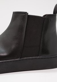 Bottine en cuir noire avec panneau latéral élastique et languette, semelle en caoutchouc plate, finition lisse et détails de couture minimaux.