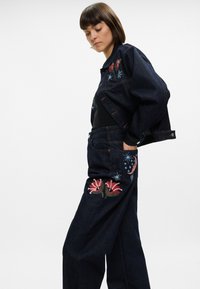 Σακάκι και παντελόνι από τζιν σε σκούρο μπλε, με λουλούδια και αστρική κεντήματα σε ζωντανά χρώματα στις τσέπες και στους ώμους.