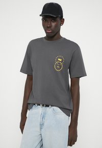 Camiseta de algodón gris con un diseño gráfico amarillo en el lado izquierdo del pecho, combinada con jeans de mezclilla azul claro y una gorra negra.