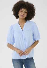 Blusa azzurro chiaro con apertura a bottoni, maniche corte a sbuffo e vestibilità rilassata. Realizzata in tessuto morbido con una texture sottile e cuciture su misura.