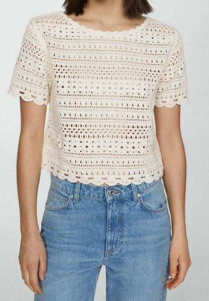 Mujer que lleva una blusa blanca de crochet de manga corta con bordes en forma de conchas y vaqueros azules de talle alto.