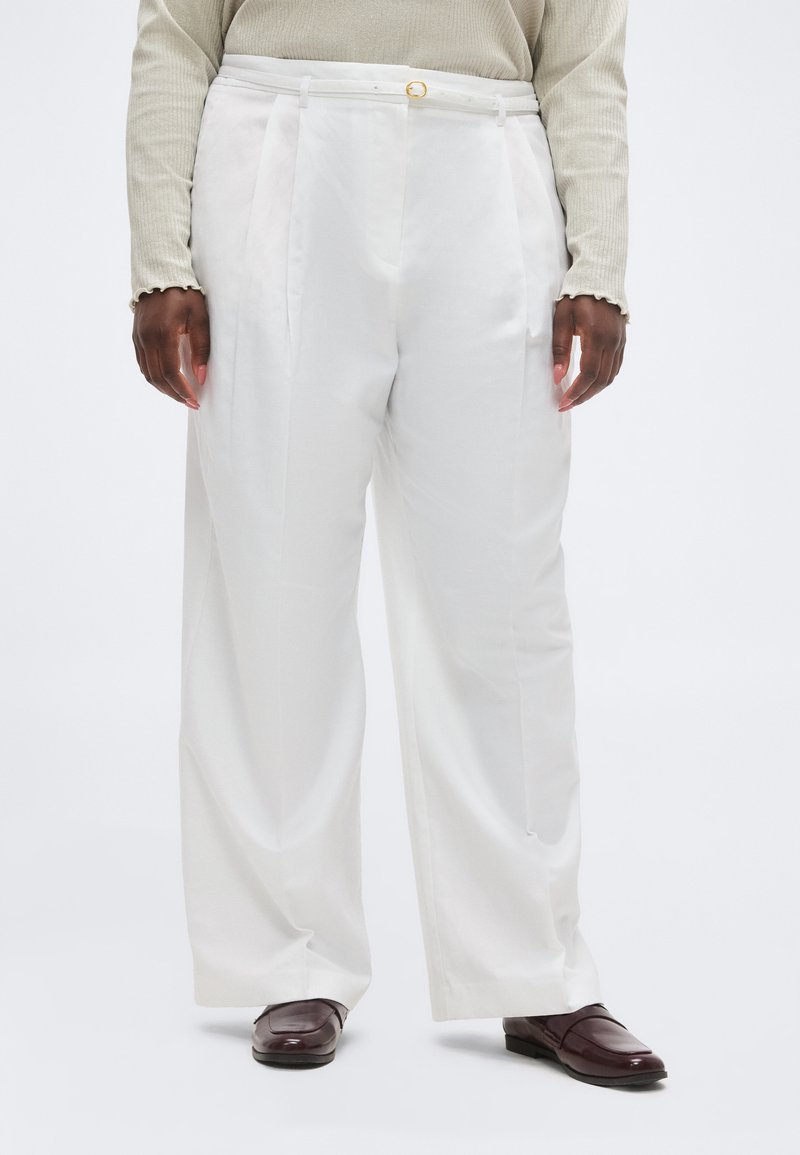 Άτομο που φοράει ψηλόμεσα λευκά παντελόνια με φαρδιά πόδια, ένα μπεζ μακρυμάνικο τοπ και σκούρα καφέ μοκασίνια, στέκεται μπροστά σε ένα απλό φόντο.