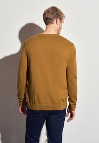 Senfgelber Pullover aus weichem Strickmaterial, mit rundem Halsausschnitt und gerippten Bündchen, kombiniert mit dunkelblauen Jeans.