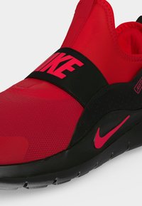 Κοντινό πλάνο κόκκινου slip-on σνίκερ Nike με μαύρο λουράκι που γράφει NIKE με ροζ, μαύρη σόλα και ροζ λογότυπο Nike (swoosh).