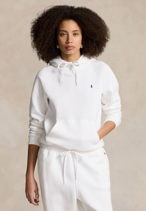 Femme aux cheveux bouclés portant un sweat à capuche blanc et un pantalon de survêtement blanc assorti, mains dans les poches, fond neutre.