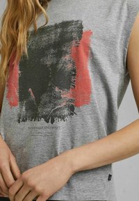 Grijze mouwloze t-shirt met een gestructureerd grafisch ontwerp in zwart en rood, met een bedrukte label aan de onderzijde links en een merketiket aan de zijkant.