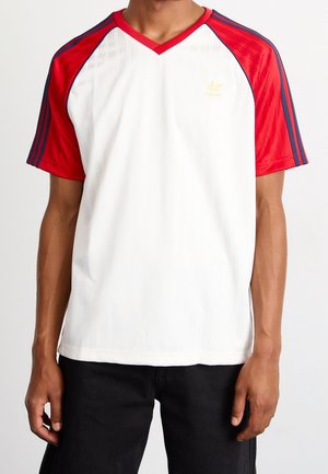 Biała sportowa koszulka z czerwonymi krótkimi rękawami, niebieskimi paskami i dekoltem w kształcie litery V. Posiada subtelną fakturę; złote logo Adidas na piersi.
