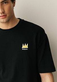 Fiatal férfi, fekete pólóban, amelyen kis sárga korona és "BASQUIAT" felirat látható a mellkasán, sima világos fal előtt áll.