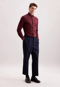 Camicia bordeaux con bottoni frontali e colletto a punta, abbinata a pantaloni plissettati blu navy e scarpe di pelle nere. Tessuto liscio, vestibilità su misura.