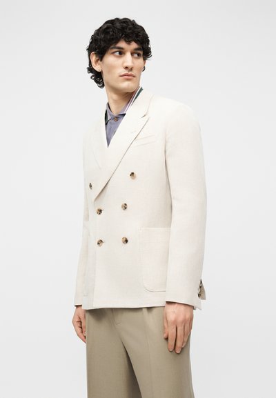 Jeune homme aux cheveux bouclés et foncés portant un blazer crème à double boutonnage, une chemise bleue à col et un pantalon beige, regardant vers sa gauche.