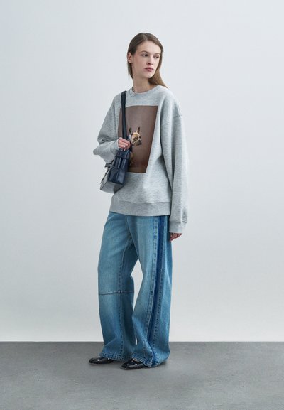 Γκρι υπερμεγέθη μπλούζα φούτερ με μεγάλο τύπωμα σκύλου στο στήθος, σε συνδυασμό με τζιν με φαρδιά πατζάκια και ρίγες στο πλάι, και μαύρα παπούτσια.