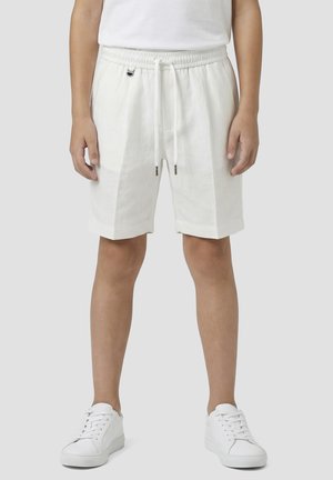 Persona con pantalones cortos blancos con cordón, zapatillas blancas y una camisa blanca, de pie frente a un fondo gris claro liso.