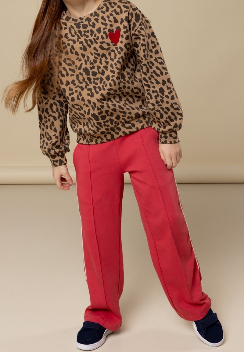 Enfant portant un t-shirt à manches longues imprimé léopard avec un cœur rouge et un pantalon rouge, debout devant un fond beige.