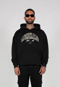 Sort hættetrøje med en frontlomme, der har et cremefarvet grafisk logo "MJ GONZALES" og en signatur. Båret med sorte solbriller og cargo-bukser.
