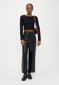 Jovem mulher vestindo um top preto cropped de manga comprida, calças largas e texturizadas de cintura alta pretas, botas pretas e uma bolsa castanha ao ombro.