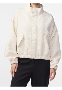 Kremowa pikowana kurtka z wysokim kołnierzem, elastycznymi mankietami i bocznymi kieszeniami. Gładka tekstura i krótki krój przyczyniają się do jej nowoczesnego wyglądu.