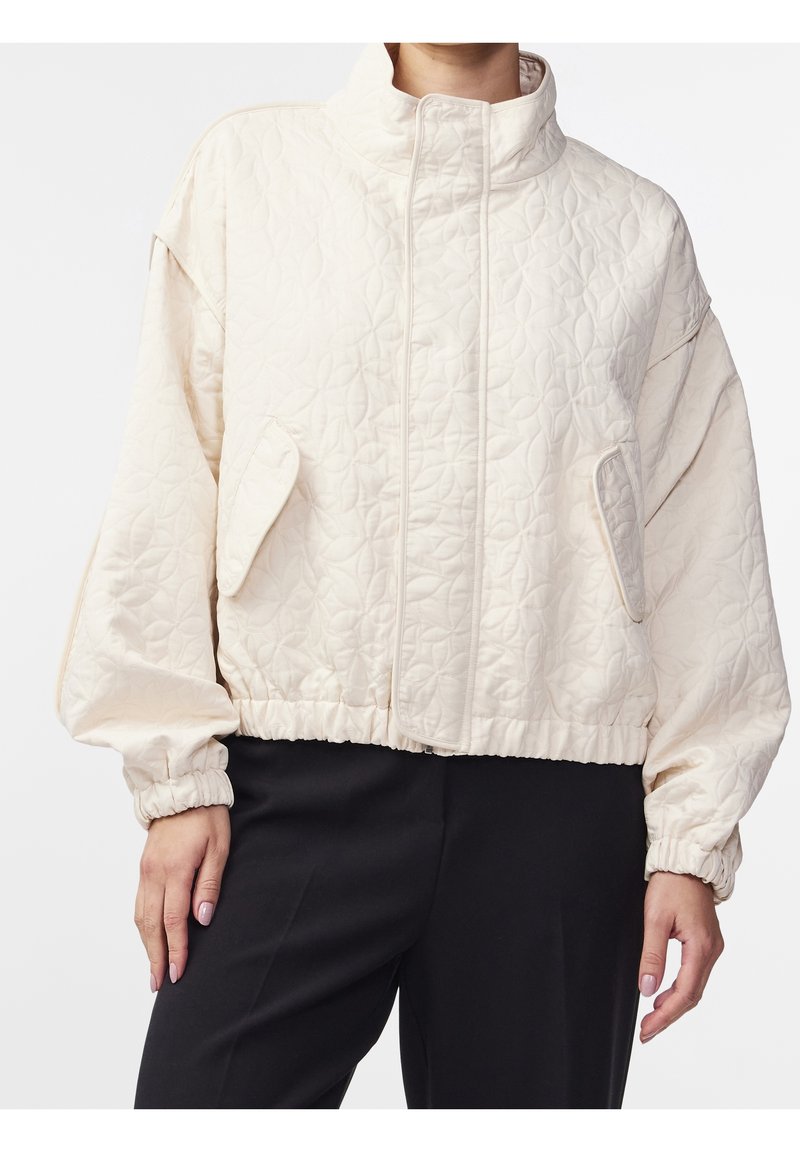 Kremowa pikowana kurtka z wysokim kołnierzem, elastycznymi mankietami i bocznymi kieszeniami. Gładka tekstura i krótki krój przyczyniają się do jej nowoczesnego wyglądu.