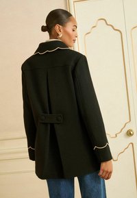 Zwarte wollen jas met een geschulpte kraag, gedetailleerde crèmekleurige bies, knoopaccenten op de rug en een losse pasvorm. Gestyled met denim.