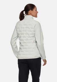 Ljusgrå mid-lagerjacka med en puffy bak, slät front, hög krage och subtil logotypdetalj. Har långa ärmar och en figurnära design.