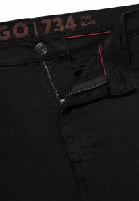 Black denim jeans featuring a zip fly with a gold zipper, silver button, and a red accent inside the waistband. Label reads "GO 734 STAY BLACK."