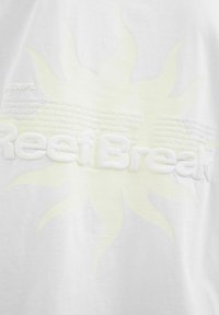 Hvid T-shirt med et struktureret soldesign i lys gul med præget tekst "ReefBreak" og yderligere tekstmønstre nedenfor. Blødt bomuldsstof.