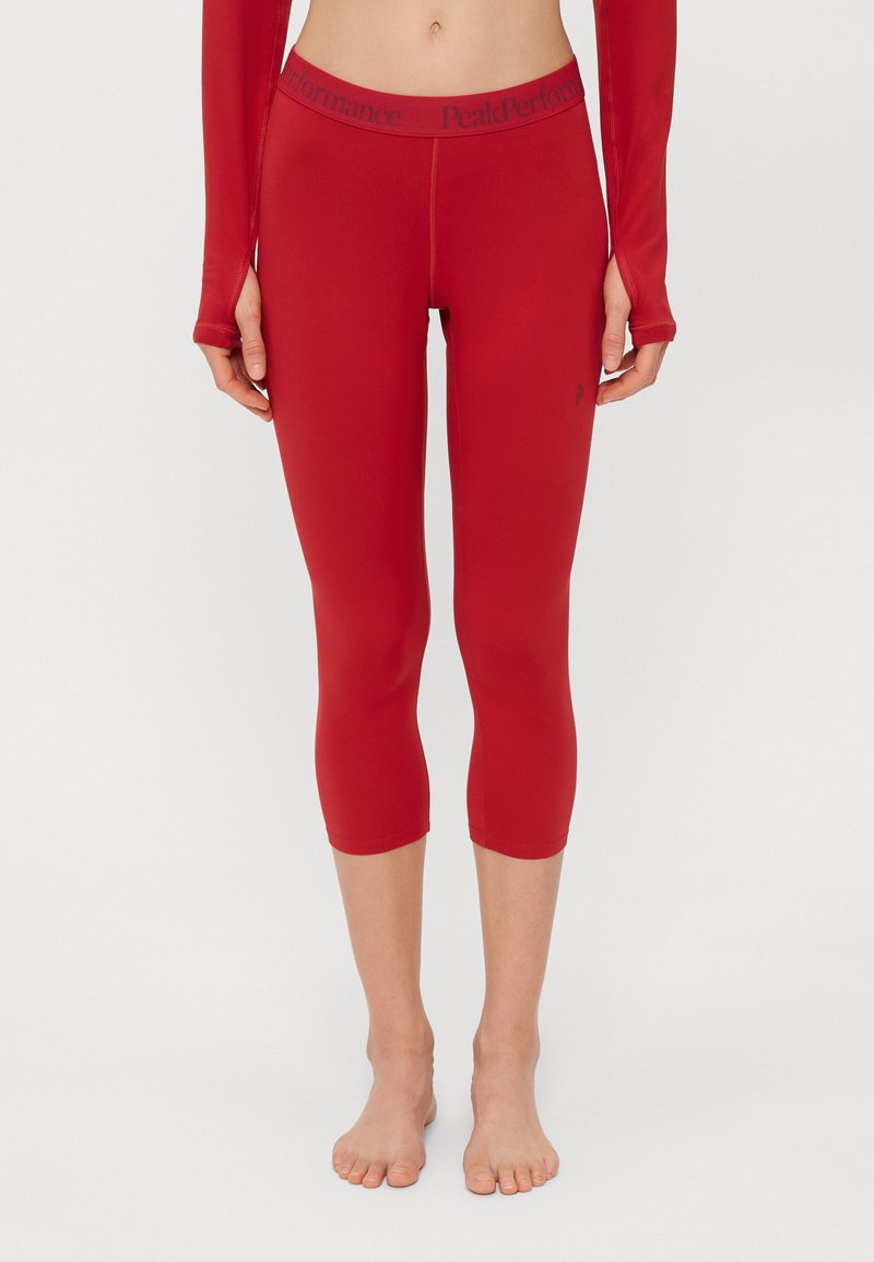Leggings ativos vermelhos com um design ajustado e comprimento três quartos, apresentando um logo da Peak Performance na cintura e um tecido liso e elástico.