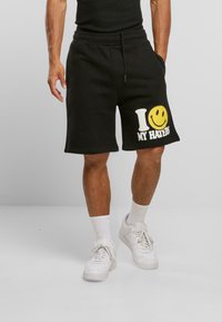 Svarte bomullsshorts med en avslappet passform, med et hvitt og gult grafisk trykk som leser "I ❤️ MY HATERS" på venstre ben, sammen med hvite sneakers.