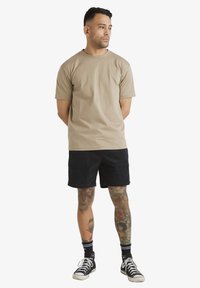 T-shirt en coton beige à manches courtes, associé à un short en velours côtelé noir. Baskets montantes noires et tatouages colorés visibles sur les jambes.