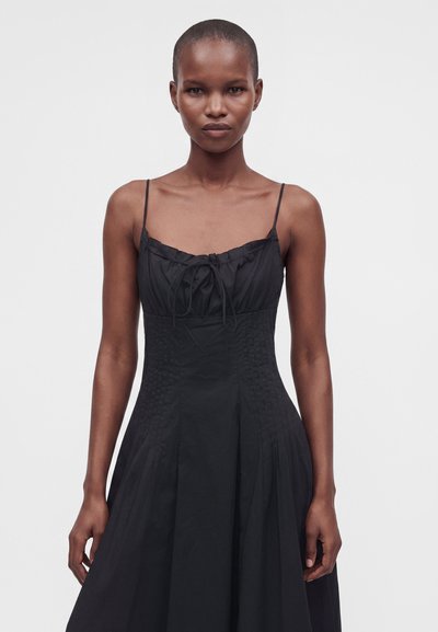 Femme aux cheveux courts portant une robe noire sans manches avec des fines bretelles et un détail nœud sur la poitrine, debout devant un fond uni.