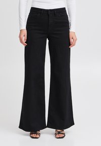 Donna che indossa jeans neri a vita alta e a gamba larga abbinati a una maglietta bianca a maniche lunghe e sandali neri con cinturini su uno sfondo bianco.