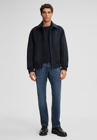 Granatowa kurtka zapinana na zamek, noszona na ciemnej koszulce, w połączeniu z niebieskimi dżinsami i czarnymi butami. Kurtka ma wysoki kołnierz oraz boczne kieszenie.