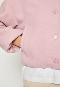 Ružová textúrovaná bunda s veľkými gombíkmi a prednou kapsou. Nosená cez biele tričko, predvádzajúca voľný strih a manžety na rukávoch.