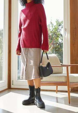 Person wearing red cable knit sweater, checked midi skirt, black chunky boots, holding black handbag, standing in sunlit room near chair and window.