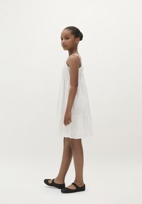 Weißes Baumwollkleid mit gestuften Design, bestickten Details, dünnen Trägern und lockerer Passform. Trägt man mit schwarzen Ballettschuhen.
