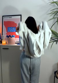 Hellgrauer Baumwollhoodie mit Kängurutasche und lockerer Passform, der vor einer Kulisse dekorativer Gegenstände angezogen wird.