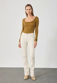 Γυναίκα που φοράει ένα εφαρμοστό ελαιώδη μπλουζάκι με στρογγυλή λαιμόκοψη και μακριά μανίκια, ψηλόμεσες κρέμα παντελόνες, μπεζ μποτάκια με μυτερή μύτη, στέκεται μπροστά σε απλό φόντο.