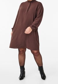 Hnědé šaty s dlouhým rukávem, volným střihem a volánkovým límcem, zapínání na knoflíky. Nosené s černými kotníkovými botami a průhlednými punčochami.