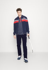 Marine sportjacket met rode en witte strepen, gestreepte shirt, marineblauwe broek en blauwe sneakers, met een tennisracket in de hand.