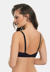 Donna con lunghi capelli castani che indossa un reggiseno nero, vista di spalle, che guarda oltre la spalla su uno sfondo bianco semplice.