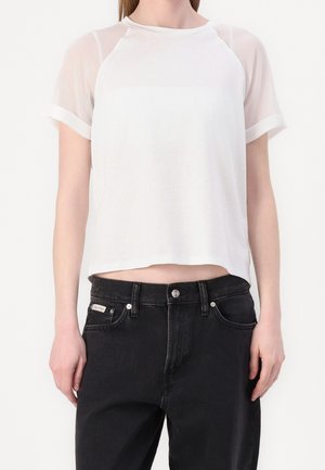Donna che indossa un top corto bianco a maniche corte con tessuto trasparente sulle maniche e jeans neri a vita alta.