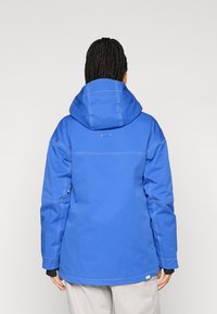 Μπλε αδιάβροχο μπουφάν με κουκούλα, που διαθέτει αντίθετα λευκά ραφάκια, χαλαρή εφαρμογή και ελαστικές μανσέτες. Λείος υφασμάτινος θρόισμα, χωρίς ορατά σχέδια.