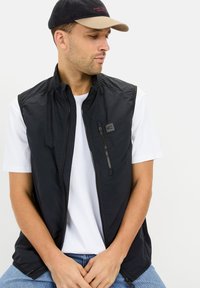 Schwarze ärmellose Weste mit hohem Kragen, Reißverschluss vorne und Taschen auf der linken Brust; getragen über einem weißen T-Shirt und blauen Jeans.