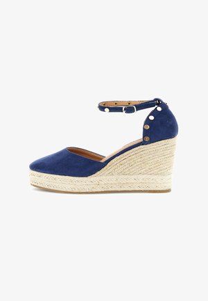Chaussure compensée en daim bleu marine avec plateforme en jute tressé, bride de cheville avec boucle, et clous métalliques décoratifs sur la zone du talon.