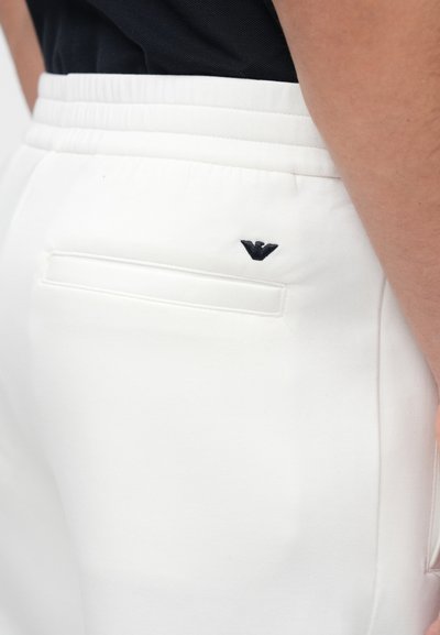 Biele teplákové nohavice s elastickým pásom, s vreckom vzadu a malým čiernym logom. Hladká textúra látky, priliehavý strih.