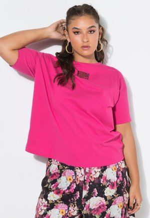 Vrouw met lang golvend haar draagt grote oorringen, een felroze T-shirt en bloemige broek, poserend met één hand achter haar hoofd.